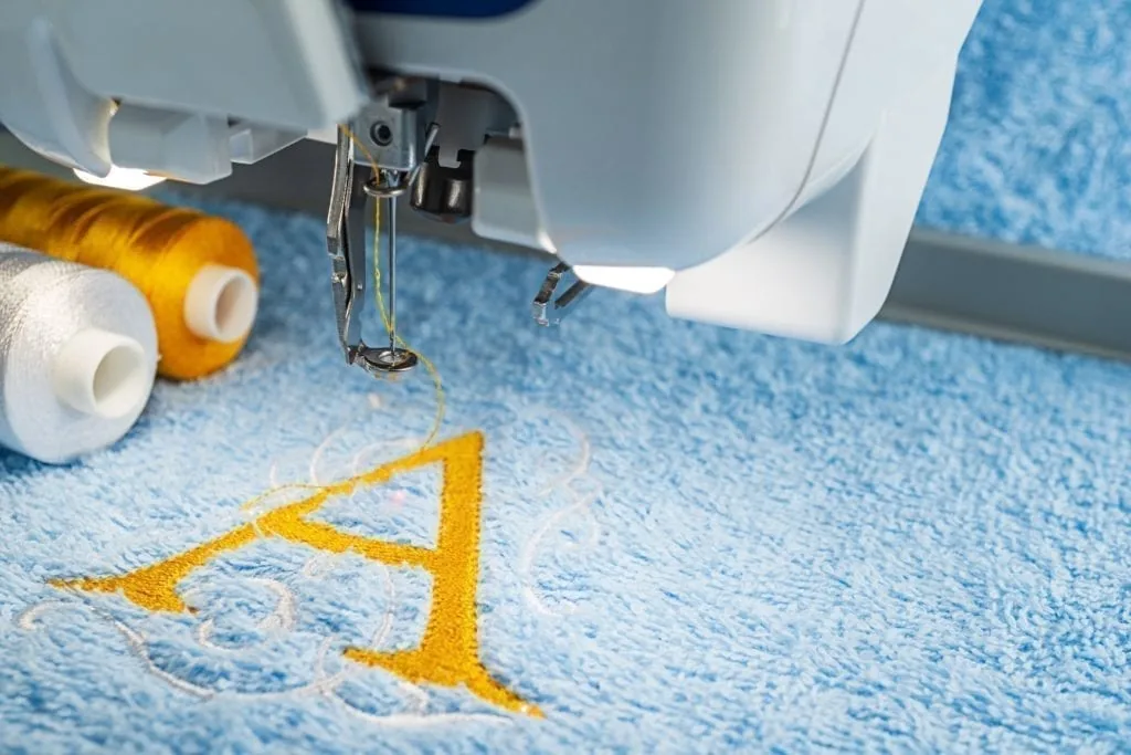 Stickmaschine stickt einen goldenen Buchstaben in ein hellblaues Frottiertuch